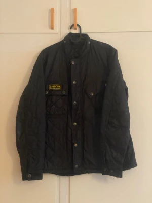 Svart quiltad jacka från Barbour - Säljer en svart quiltad jacka från Barbour med tryckknappar och hög krage. Jackan har Barbour International-märke på bröstet, två bröstfickor med knapp och lång ärm. Perfekt för dig som gillar klassisk och stilren design.