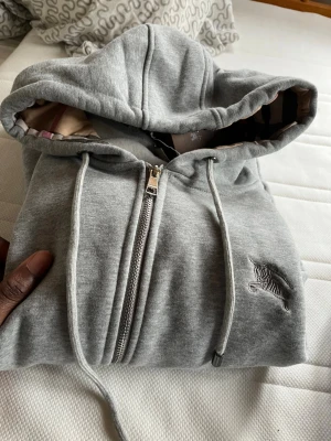 Grå zip-hoodie från Burberry - Snygg grå zip-hoodie från Burberry med klassiskt rutigt foder i huvan och broderad logga på bröstet. Mjuk bomullsblandning, dragsko i huvan och tvåvägsdragkedja framtill. Perfekt för en avslappnad men stilren look.