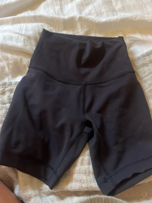 Svarta cykelshorts Lululemon - Säljer ett par svarta cykelshorts från Lululemon med hög midja och tight passform. Perfekta för träning eller chill dagar. Tillverkade i ett stretchigt syntetmaterial som sitter skönt och följer kroppen. Användt två gånger