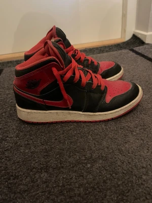 Air Jordan 1  - Säljer ett par Air Jordan 1 Mid i klassisk svart och röd färg. Skorna har läderöverdel, röda snören och ikoniska Jordan-loggor på sidan och plösen. Perfekta för dig som gillar streetwear och vill sticka ut med en tidlös sneaker.