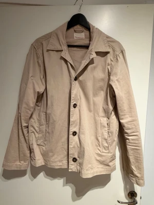 Beige overshirt från Knowledge Cotton Apparel - Stilren beige overshirt från Knowledge Cotton Apparel med klassisk krage, knäppning framtill och två sidofickor. Tillverkad i ett mjukt bomullstyg som ger en avslappnad känsla. Perfekt lager-på-lager-plagg för en clean look.           Nypris runt 1600kr