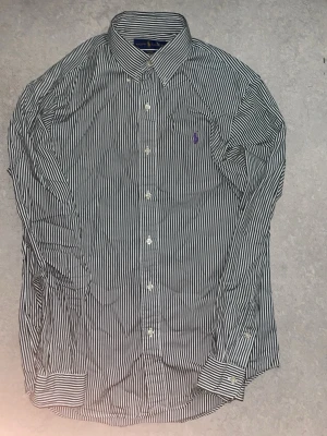 Randig skjorta från Ralph Lauren - Klassisk långärmad skjorta från Ralph Lauren med mörkgröna vita ränder och broderad lila logga på bröstet. Skjortan har knappar framtill och vid ärmsluten. Perfekt för en stilren och tidlös look.