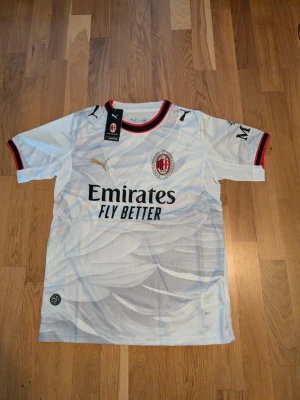 AC Milan vit bortatröja Puma. Storlek. M.L - Säljer en vit AC Milan bortatröja från Puma med klubbmärke och sponsortryck på bröstet. Tröjan har svarta och röda detaljer vid krage och ärmslut samt guld Puma-logga. Tillverkad i lätt och ventilerande material, perfekt för fotbollsträning.