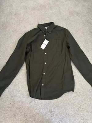 Helt ny oanvänd grön linneskjorta från Jack & Jones - Helt ny oanvänd stilren grön skjorta från Jack & Jones i linne med klassisk krage och vita knappar. Skjortan har lång ärm och en bröstficka, perfekt för en avslappnad men snygg look. Enkel att matcha med jeans eller chinos.