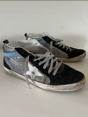 Glittriga GGDB Mid Star sneakers - Snygga Golden Goose Mid Star sneakers med svart mocka, silverglittriga paneler, vit stjärndetalj och ljusblå rand. Klassisk GGDB-design med slitna detaljer och vita skosnören. Perfekta för dig som vill sticka ut med en cool och trendig look.