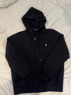 Ralph Lauren zip hopdie - Snygg hoodie med dragkedja från Polo Ralph Lauren. Klassisk design med huva och dragsnören, samt den ikoniska vita logotypen broderad på bröstet. Mjuk bomullskänsla och ribbade muddar vid ärmslut och nederkant.
