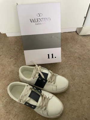 Valentino Garavani vita sneakers - Snygga vita sneakers från Valentino Garavani med bred svart rem och silvriga nitar på sidorna. Skorna har klassisk låg modell, vita snören och diskret logga på plösen. Perfekta för dig som gillar stilrena och exklusiva detaljer.