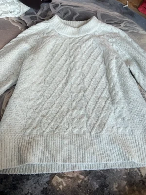 Vit kabelstickad tröja från 157 - Snygg vit kabelstickad tröja från 157 med ribbade muddar och rund hals. Tröjan har ett klassiskt flätmönster framtill och är perfekt för lager-på-lager-stil. Passar dig som gillar en enkel men trendig look.
