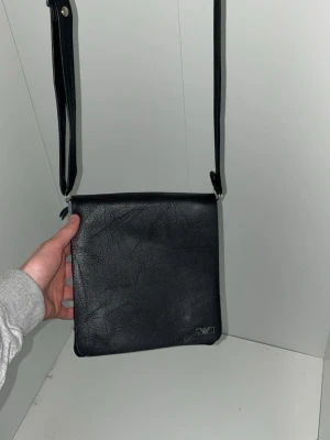 Svart axelväska i skinnimitation - Stilren svart axelväska från AXE i skinnimitation med justerbart axelband. Väskan har en fyrkantig form och en enkel design utan synliga fickor på utsidan. Perfekt för dig som gillar minimalistisk stil.