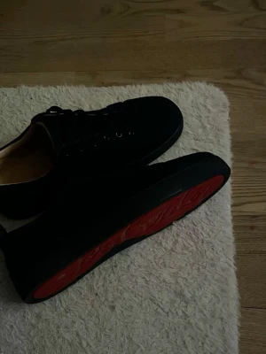 Svarta Louboutin sneakers med röd sula - Snygga svarta sneakers från Christian Louboutin med den ikoniska röda sulan. Skorna har lågt skaft, svarta skosnören och ovandel i mocka. Perfekta för dig som vill sticka ut med en exklusiv och trendig look.
