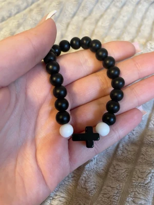 Svart och vitt pärlarmband med kors - Snyggt armband med svarta och vita pärlor samt ett svart kors i mitten. Armbandet är elastiskt och passar de flesta handleder. Perfekt accessoar för dig som gillar stilrena och trendiga smycken med en personlig touch.
