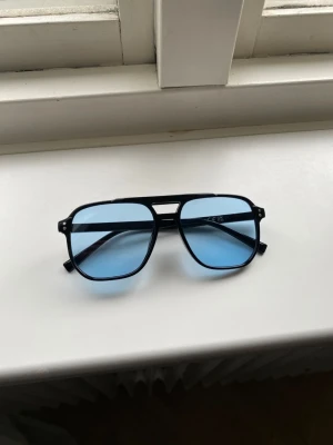 Svarta solglasögon med blåa glas - Snygga solglasögon med svart båge och rektangulär form. Glasen är ljusblå och ger en cool retrovibe. Perfekta för dig som vill sticka ut med en unik accessoar. Bågarna är i plast och har en klassisk design med dubbla bryggor.