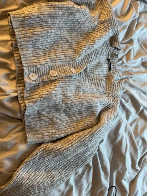 Grå ribbad kofta från Gina Tricot - Mysig grå ribbstickad kofta från Gina Tricot med v-ringning och tre stora vita knappar framtill. Kortare modell med breda muddar och lång ärm. Perfekt att slänga över en topp för en chill look.