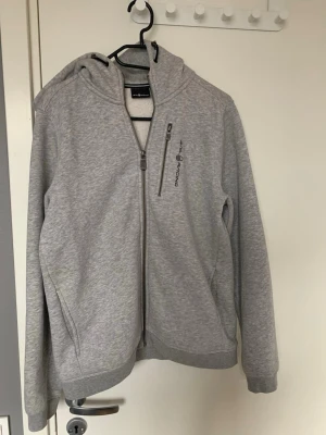 Ljusgrå Sail Racing zip-hoodie  - Ljusgrå hoodie med dragkedja från SAIL RACING. Tröjan har huva, bröstficka med dragkedja och diskret logga på bröstet samt en liten detalj på huvan. Mjuk bomullsblandning och ribbade muddar vid ärmslut och nederkant. Sail racing tröja som är riktigt skön och passar till det mesta. 