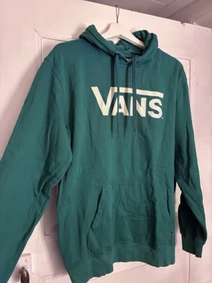 Grön hoodie från Vans med logga - Snygg grön hoodie från Vans med stor vit logga på bröstet. Klassisk känguruficka framtill och justerbar huva med dragsko. Tillverkad i mjukt bomullsmaterial som är skönt att ha på sig. Perfekt för en avslappnad streetwear-look.