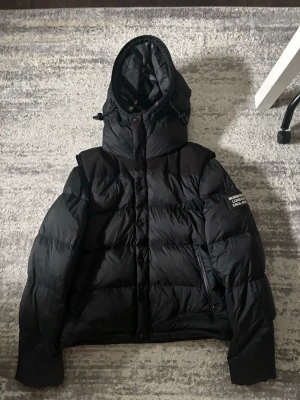 Svart pufferjacka/väst Burberry  - Säljer en svart pufferjacka från Burberry med avtagbara ärmar så den kan användas som väst. Jackan har huva, dragkedja och knappar framtill samt två fickor. Märkeslogga på ärmen och quiltad insida. Perfekt för kalla dagar och som väst i sommar 