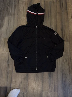 Svart Moncler vindjacka med huva - Svart vindjacka från Moncler med huva och dragkedja framtill. Jackan har Moncler-logga på ärmen och rödvit rand på huvan. Två fickor med tryckknappar och justerbar dragsko vid huvan. Perfekt för en sportig och stilren look. PASSAR M med 