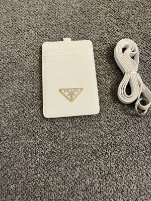 Vit korthållare från Prada - Snygg vit korthållare från Prada i läder med klassisk trekantig logga i metall framtill. Kommer med matchande axelrem som enkelt kan fästas. Perfekt för att bära kort och ID på ett stilrent sätt.