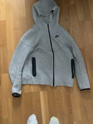 Grå hoodie från Nike med dragkedja - Snygg grå hoodie från Nike med svart logga på bröstet och hel dragkedja framtill. Två svarta dragkedjeförsedda fickor på sidorna och svarta detaljer vid ärmsluten. Tillverkad i mjukt sweatshirtmaterial och har en klassisk huva.