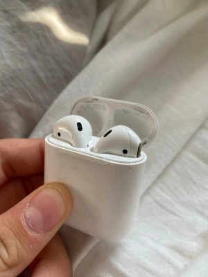 Apple AirPods med laddningsetui - Säljer ett par vita Apple AirPods med tillhörande laddningsetui. Trådlösa hörlurar med stilren och kompakt design i blank plast. Perfekta för musik, poddar och samtal på språng. Laddningsetuiet är smidigt att ta med och håller lurarna laddade hela dagen.