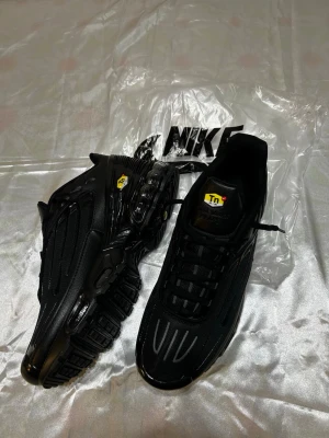 Nike Air Max Plus Tn helsvarta sneakers - Säljer ett par helsvarta Nike Air Max Plus Tn sneakers med klassisk Tn-logo på sidan och tungan. Skorna har en sportig siluett, synliga Air Max-enheter i sulan och ovandel i mesh och syntet. Perfekta för dig som gillar en clean och modern look.