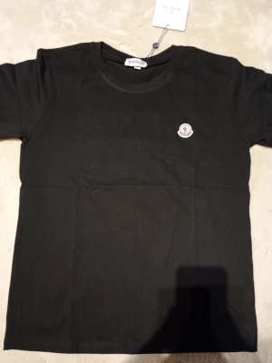Svart t-shirt från Moncler - Stilren svart t-shirt från Moncler med rund halsringning och klassisk passform. T-shirten har ett broderat Moncler-märke på bröstet och är tillverkad i mjuk bomull som känns skön mot huden. Perfekt basplagg för en clean och enkel look.