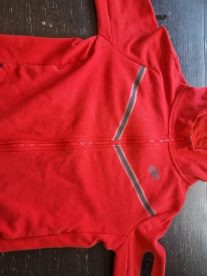 Röd Nike hoodie med dragkedja - Snygg röd hoodie från Nike med hel dragkedja framtill och huva. Har grå och svarta ränder diagonalt över bröstet samt Nike-logga på vänster sida. Tillverkad i mjukt bomullsmaterial, perfekt för en sportig och avslappnad stil. Dock kan dragkedjan fastna ibland men är tar 3 sekunder att fixa till.