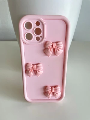 Rosa mobilskal med rosetter till iPhone 12 Pro  - Sött ljusrosa mobilskal med tre upphöjda rosetter på baksidan. Skalets material är mjukt silikon som ger bra grepp och skydd. Perfekt för dig som vill ha en gullig och trendig look till mobilen.