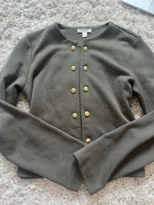 Olivgrön kofta med guldknappar H&M - Snygg olivgrön kofta från H&M med dekorativa guldknappar i två rader framtill. Koftan har rund halsringning och långa ärmar. Perfekt för att lyfta en enkel outfit med lite extra detaljer.