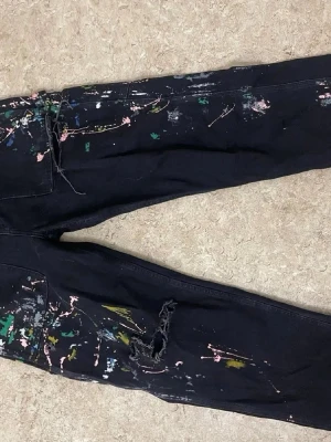 Svarta jeans med färgstänk och slitningar - Unika svarta jeans med färgstänk i grönt, rosa, vitt och gult. Jeansen har en avslappnad passform och slitningar vid knät för en edgy look. Perfekta för dig som vill sticka ut med en kreativ och konstnärlig stil.