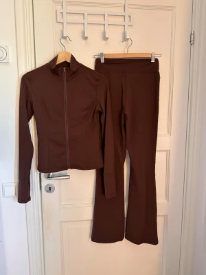 Brunt loungewear-set med ziptröja - Snyggt brunt loungewear-set med figurnära ziptröja och matchande utsvängda byxor. Tröjan har hög krage och dragkedja framtill. Mjukt och stretchigt material som passar perfekt för chill dagar hemma men funkar även som gymkläder. 