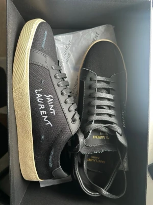 Svarta Saint Laurent sneakers - Svarta sneakers från Saint Laurent med vit logga på sidan. Skorna har svart canvas och läderdetaljer, samt en beige sula som ger en snygg kontrast. Klassisk låg modell med svarta skosnören och diskreta guldiga detaljer på insidan.
