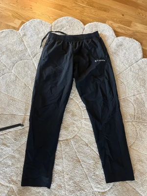 Svarta träningsbyxor från GTsch - Svarta träningsbyxor från GTsch med resår och dragsko i midjan. Byxorna har en rak passform och är tillverkade i ett lätt, funktionsmaterial som passar perfekt för sport och träning. Diskret logga på ena benet.
