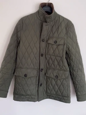 Olivgrön quiltad jacka från Morris - Snygg olivgrön quiltad jacka från Morris med hög krage och fyra framfickor med knappar. Jackan har ett rutigt mönster, långa ärmar och stängs med knappar framtill. Perfekt för dig som gillar klassisk och stilren design.