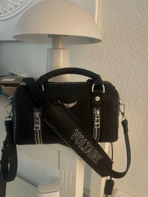  Zadig & Voltaire handväska -  svart väska från Zadig & Voltaire i  läder med silvriga dragkedjor och detaljer. Väskan har ett brett axelband med texten VOLTAIRE och ett litet emblem framtill. Den är i bra skick och man kan välja om man antigen vill ha banden på eller avtagna, det medföljd dustbag till väskan, väskan är köpt här på Plick sen tidigare!🤗 säljer eftersom jag skulle vilja testa något nytt