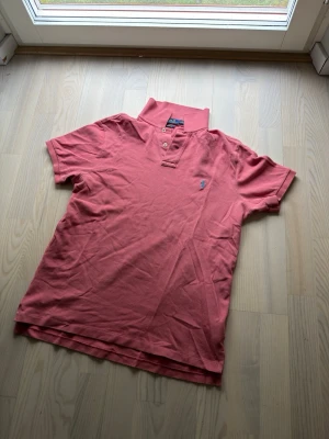 Rosa pikétröja från Polo Ralph Lauren - Säljer en klassisk rosa pikétröja från Polo Ralph Lauren med korta ärmar och blå broderad logga på bröstet. Tröjan har krage och två knappar vid halsen. Perfekt till jeans eller shorts för en avslappnad men snygg look.