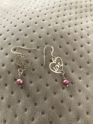 Hjärnformade örhängen med pärlor - Säljer ett par söta örhängen i silverfärgad metall med hjärtformade hängen där det står 'LOVE'. Under hjärtat hänger en lila pärla som ger en extra touch. Perfekta för dig som gillar romantiska detaljer och vill sticka ut lite extra.