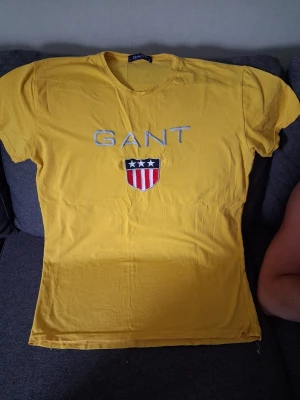 Gul GANT t-shirt med tryck - Gul t-shirt från GANT med broderad logga och ett sköldmärke med stjärnor och ränder på bröstet. Klassisk rund halsringning och korta ärmar. Perfekt för dig som gillar stilrena och sportiga plagg.