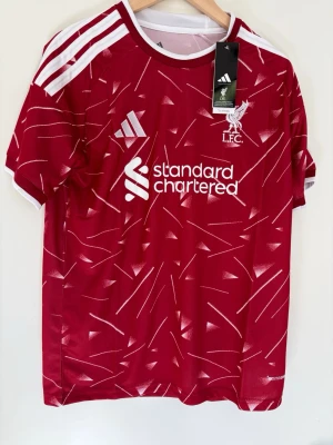 Liverpool matchtröja Adidas röd M - Liverpool FC matchtröja från Adidas i rött med vita detaljer och grafiskt mönster. Tröjan har klubbmärke, sponsorlogga och klassiska Adidas-ränder på axlarna. Tillverkad i lätt och ventilerande funktionsmaterial, perfekt för träning eller match.