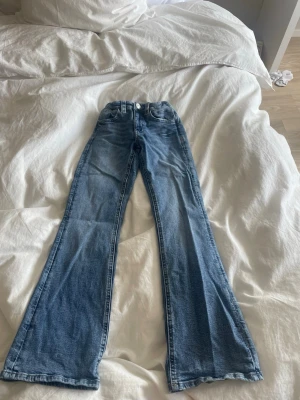 Blå bootcut jeans med hög midja - Säljer ett par ljusblå bootcut jeans med hög midja och klassisk femficksdesign. Jeansen har utsvängda ben och stängs med knapp och dragkedja. Perfekta för dig som gillar retrovibbar och en avslappnad look.