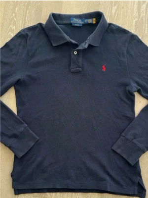 Mörkblå långärmad piké från Polo Ralph Lauren - Klassisk mörkblå långärmad pikétröja från Polo Ralph Lauren med röd broderad logga på bröstet. Skjortan har krage, knappar vid halsen och är tillverkad i mjuk bomull. Perfekt för en stilren och avslappnad look.