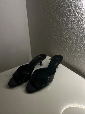 Svarta lackpumps från Mango - Säljer ett par stilrena svarta pumps från Mango i glansigt lackmaterial. Skorna har öppen tå, låg smal klack och slip-in modell som ger en trendig look. Perfekta för dig som gillar minimalistisk och modern stil.