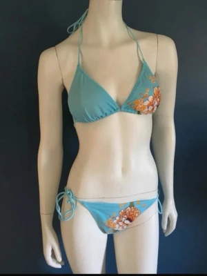Ljusblå bikini  strl 38 - Ljusblå bikini i triangelmodell med stora blommiga tryck i orange och vitt på både överdel och underdel. Knytband i nacken och på sidorna för justerbar passform. Tillverkad i mjukt syntetmaterial som torkar snabbt, perfekt för strand och pool.