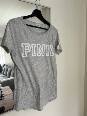 Grå T-shirt från Victorias secret PINK med tryck - Gråmelerad T-shirt från Victorias secret, PINK med stort vitt tryck framtill och detaljer på ärmen där det står 'LOVE PINK' och två vita ränder. Klassisk rund hals och korta ärmar. Perfekt för en avslappnad och sportig look.