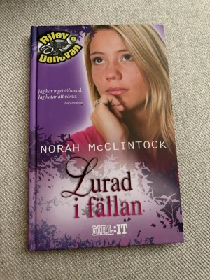 Lurad i fällan av Norah McClintock - En spännande ungdomsroman med lila hård pärm, skriven av Norah McClintock. Boken handlar om Riley Donovan och är en del av GIRL:IT-serien. Omslaget har en tonårstjej och grafiska detaljer i lila och vitt.
