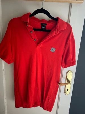 Röd pikétröja från BOSS - Snygg röd pikétröja från BOSS med klassisk krage och knappar framtill. Tröjan har korta ärmar och en liten BOSS-logga på bröstet. Slim fit-modell som passar perfekt till jeans eller shorts.