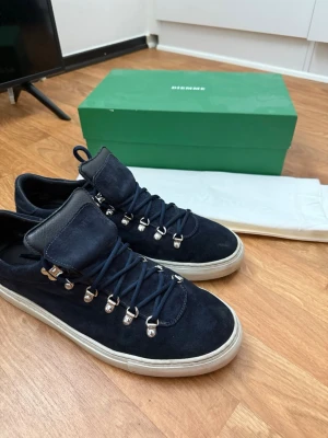 Svarta sneakers från Diemme i mocka - Snygga svarta sneakers från Diemme i mjuk mocka med robust vit sula. Skorna har metallhakar för snörningen och en unik, vadderad plös som ger en cool outdoor-vibe. Perfekta för dig som gillar stilrena detaljer och premiumkänsla. Skorna tvättas innan köp med mocka borste och medel