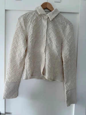 Beige skjorta med strukturmönster - Säljer en beige skjorta med zigzag-strukturmönster och klassisk krage. Skjortan har knappar framtill och långa ärmar med breda manschetter. Materialet känns lite tjockare och ger en unik look. Perfekt för dig som vill sticka ut lite extra.
