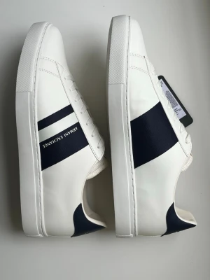 Vita Armani Exchange skor med blå detalj - Snygga vita sneakers från Armani Exchange med svart sidopanel och logga. Klassisk låg modell med vita snören och diskret branding på sidan. Tillverkade i slätt syntetmaterial för en clean look.
