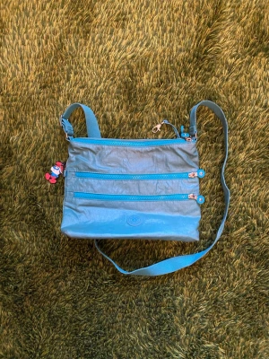 Blå axelväska från Kipling - Säljer en blå axelväska från Kipling med tre dragkedjefack framtill och justerbar axelrem. Väskan är i slitstarkt nylonmaterial och har en söt liten nyckelring. Smidig och snygg väska till vardagen!